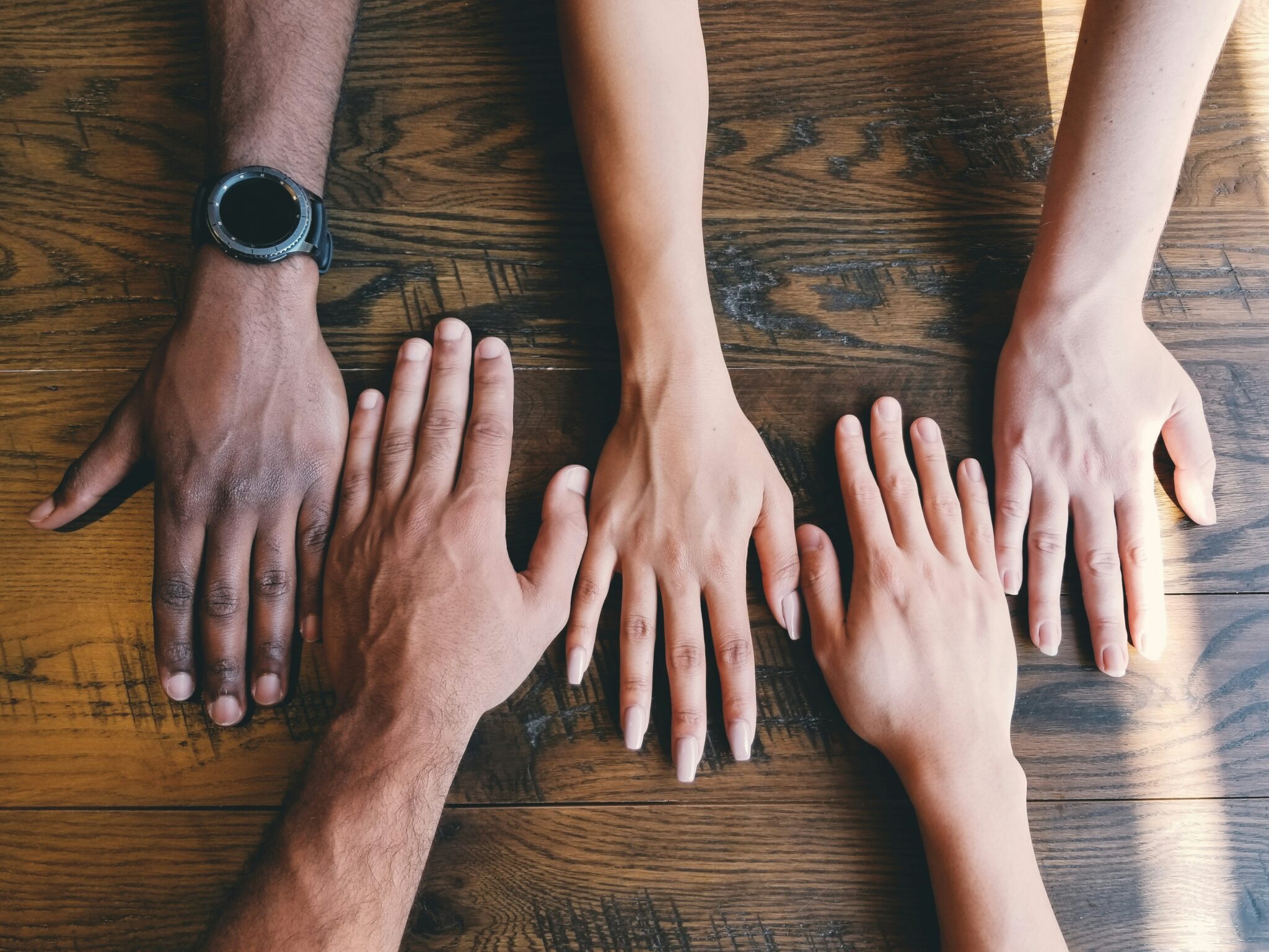 Cropped image of five hands and lower arms with varying skin tones rest on a timber table.