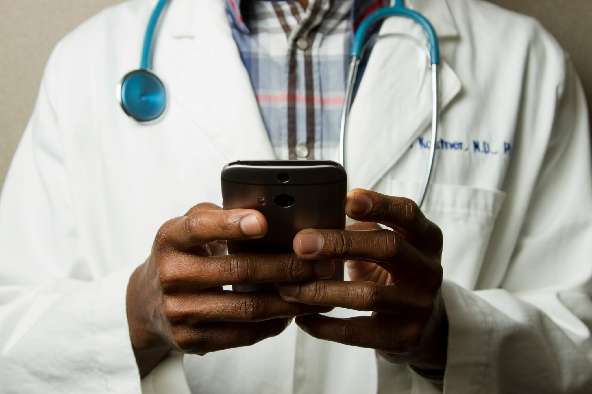 Cropped image of person holding smartphone, wearing a white coat and stethoscope.