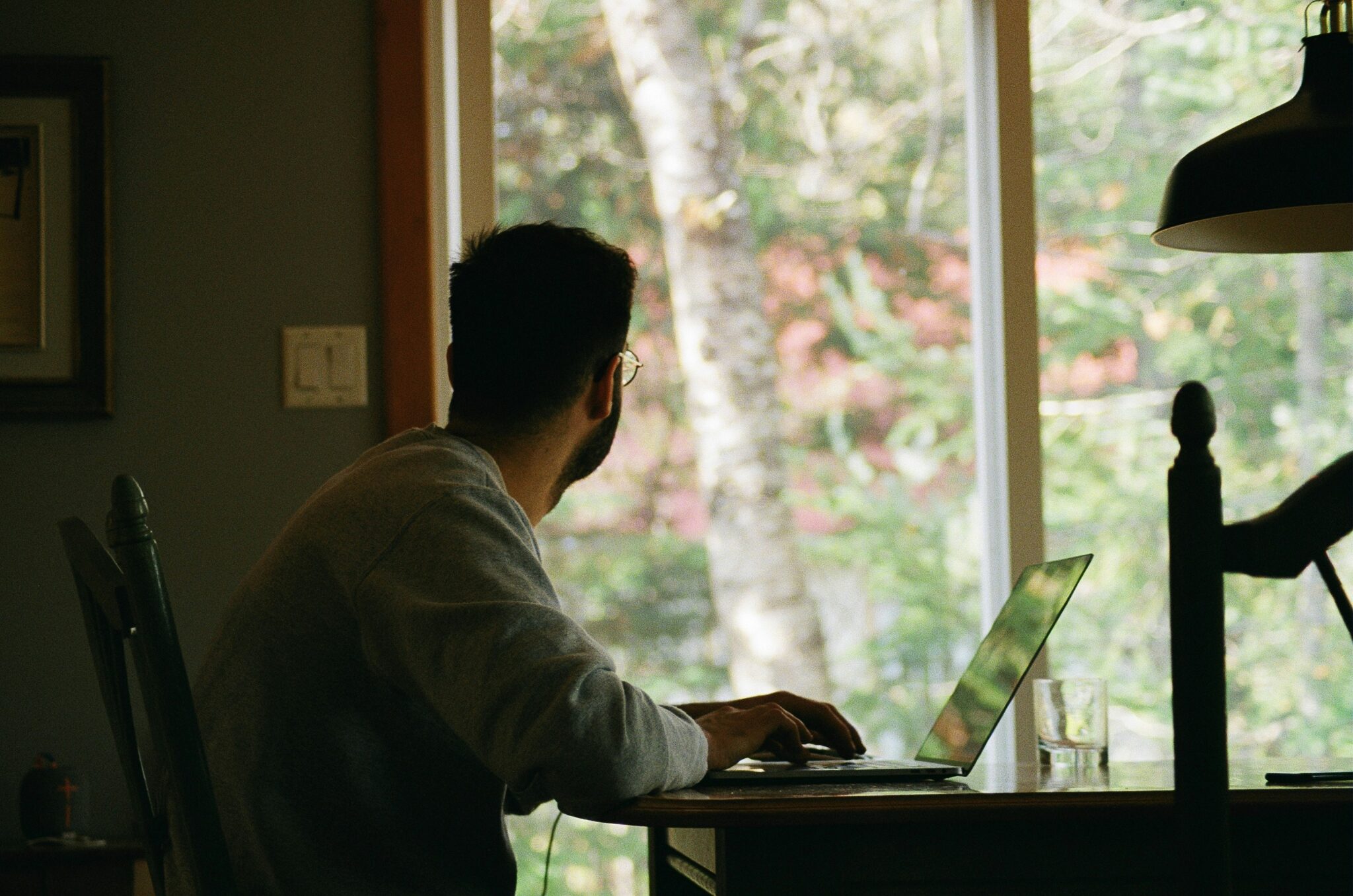 Man sits at home office desk looking out window to leafy trees outside.